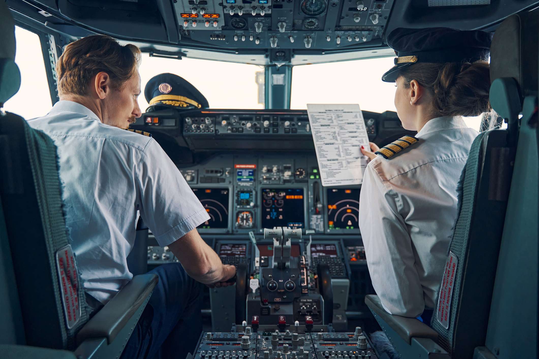 Back view of a co-pilot with a pre-flight checklist in her hand sitting by an aircraft captain in the cockpit